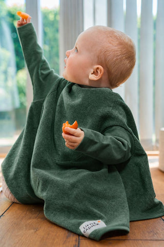 Boo Lätzchen mit Ärmeln Forest Green: Kleinkind im dunkelgrünen Lätzchen sitzt auf dem Holzboden und streckt den Arm hoch