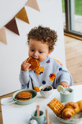 Boo Lätzchen mit Ärmeln Blueberry Pancake: Kleinkind im Lätzchen isst am Tisch mit Waffeln, hinten eine Wimpelkette