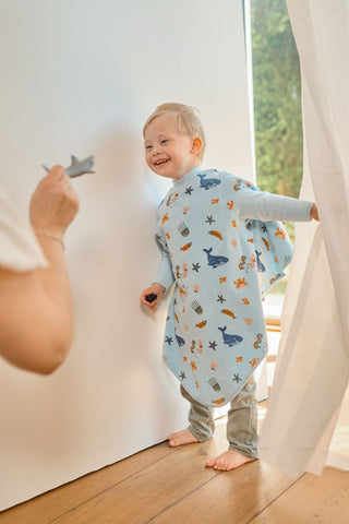 Boo Lätzchen mit Ärmeln Deap Sea: Kleinkind im Lätzchen steht an der Wand, eine Hand hält ihm ein Spielzeug hin