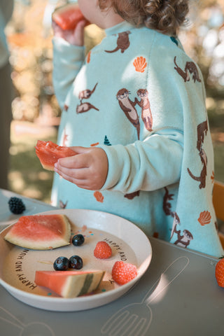 Boo Lätzchen mit Ärmeln Otter II: Kind im Lätzchen hält ein Stück Wassermelone über einem Teller mit Obst